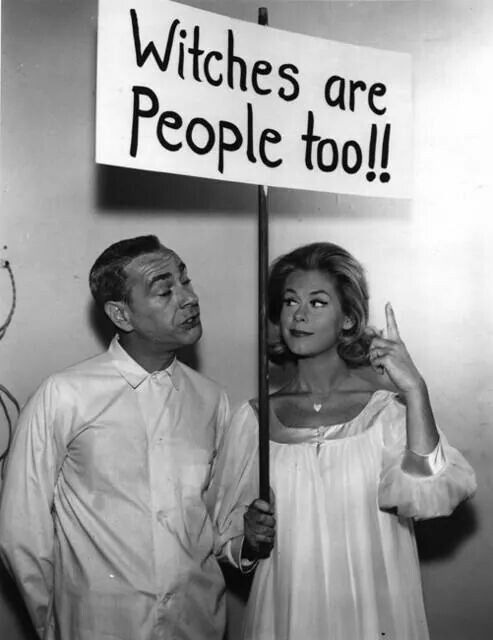 A black-and-white photo shows a man and a woman standing together, holding a sign that reads 'Witches are People too!!'. The…