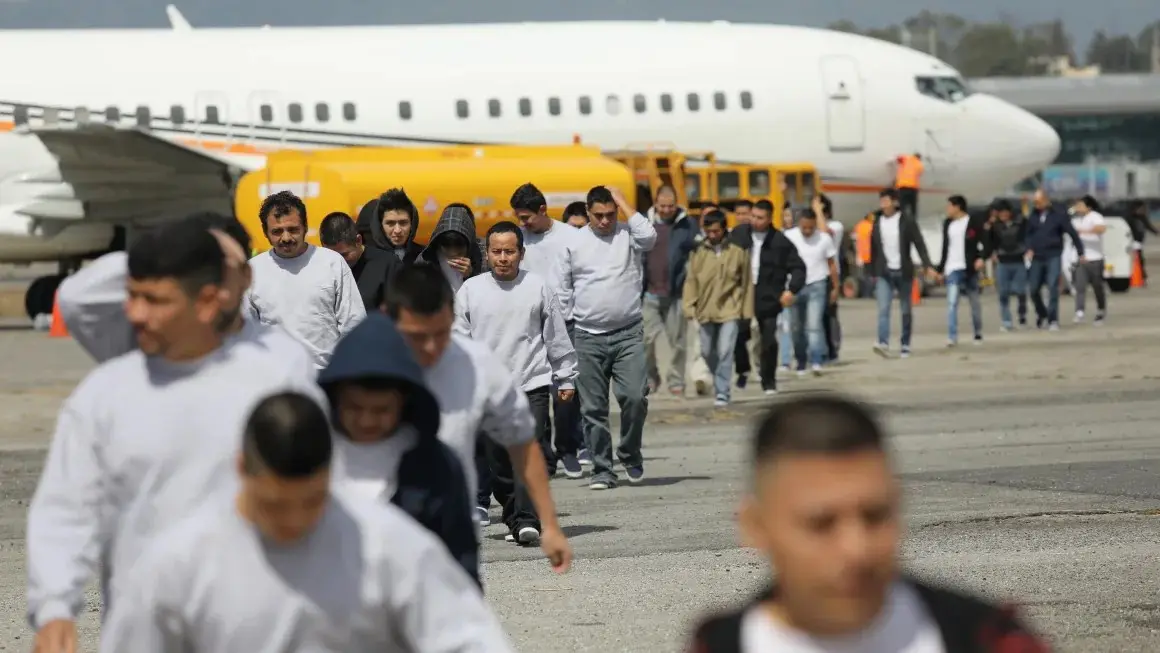 A group of men in light gray sweatshirts walk across an airport tarmac toward a large white airplane, with airport ground…