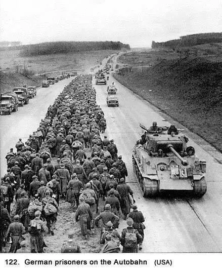 A black-and-white historical photograph shows a long column of German prisoners of war marching along a wide, paved road…