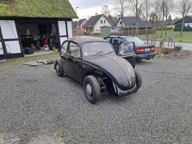 A black, vintage Volkswagen Beetle with its front wheel missing and front bumper removed, parked on gravel in front of a…