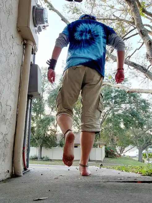 A person walks barefoot away from the camera on a concrete path beside a house, wearing a blue tie-dye shirt over a grey…