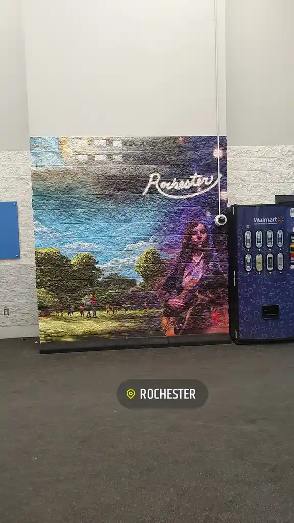 A large mural on a wall depicts a park scene with people playing and a woman playing guitar, with the word 'Rochester'…