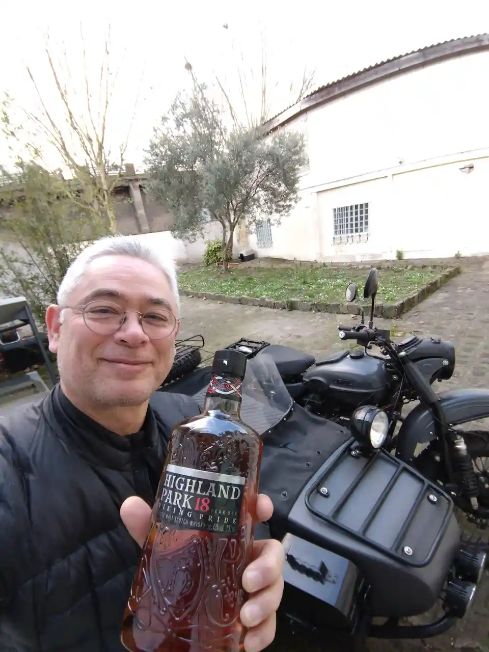 A man with white hair and glasses smiles while holding a bottle of Highland Park 18 Year Old Viking Pride whisky, standing…