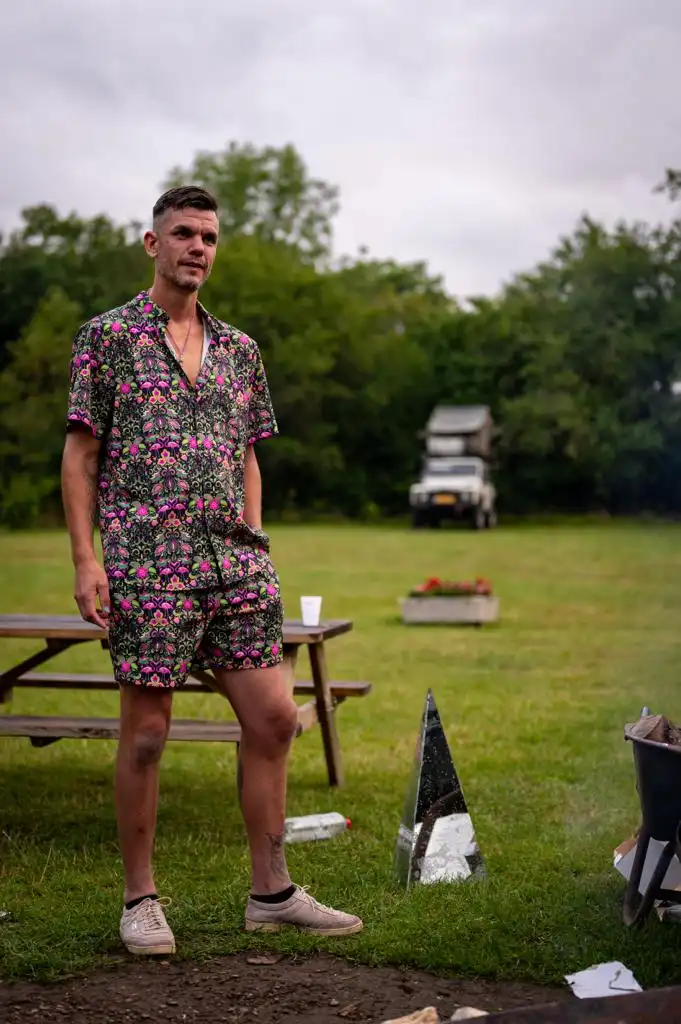 A man stands outdoors in a grassy field near a picnic table, wearing a vibrant, floral-patterned short-sleeve shirt and…