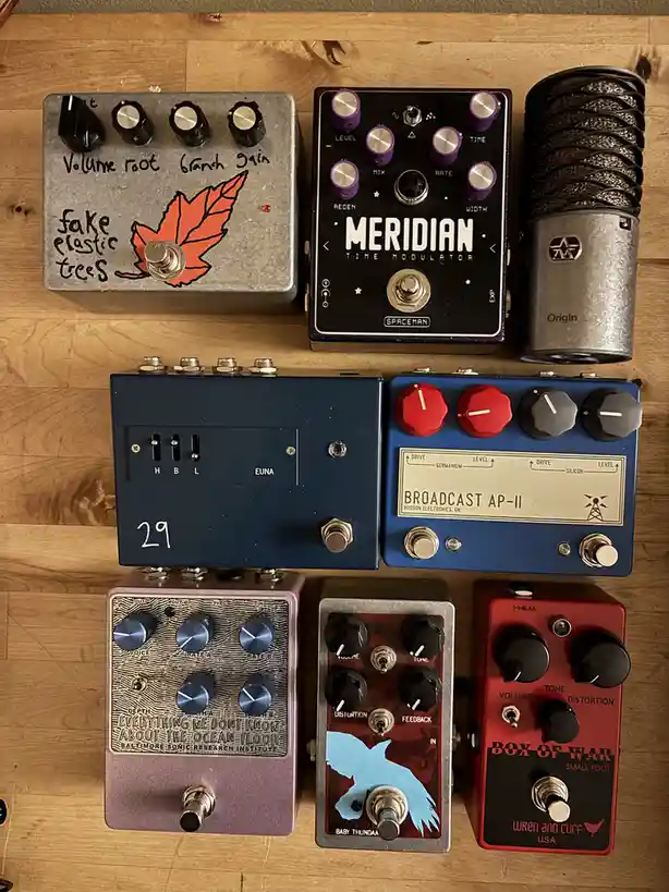 A collection of seven guitar effects pedals and a microphone arranged on a wooden surface. One pedal, a silver box with a…