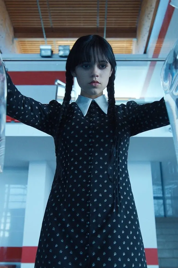 A young girl with dark pigtails and a black dress with white patterns stands with arms outstretched, looking directly at the…