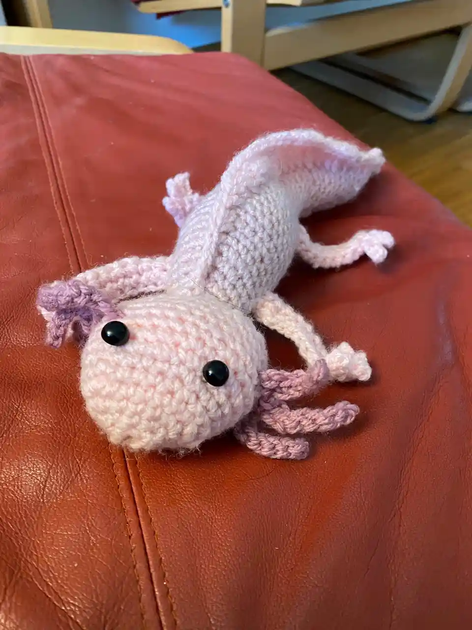 A handmade, light pink crocheted axolotl plush toy with black button eyes and purple gills lies on its side on a…