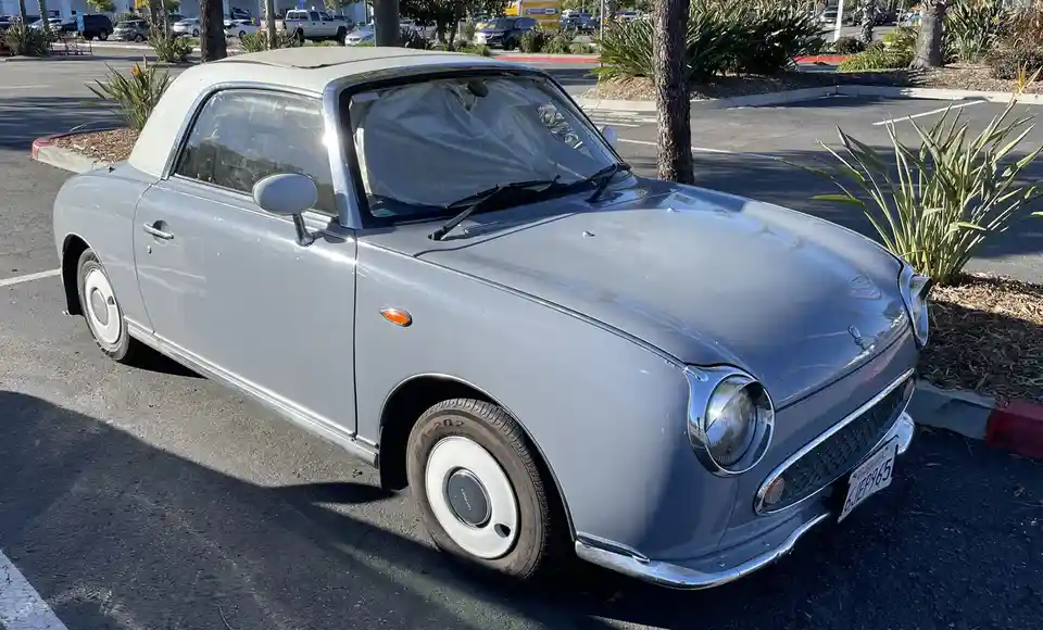 A light gray, vintage-style Nissan Figaro convertible is parked in a sunny parking lot, its windshield covered with a clear…