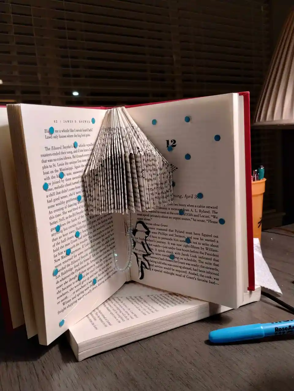 An open book with its pages folded and arranged to form the shape of a bird, with blue dots marking the folds and the number…