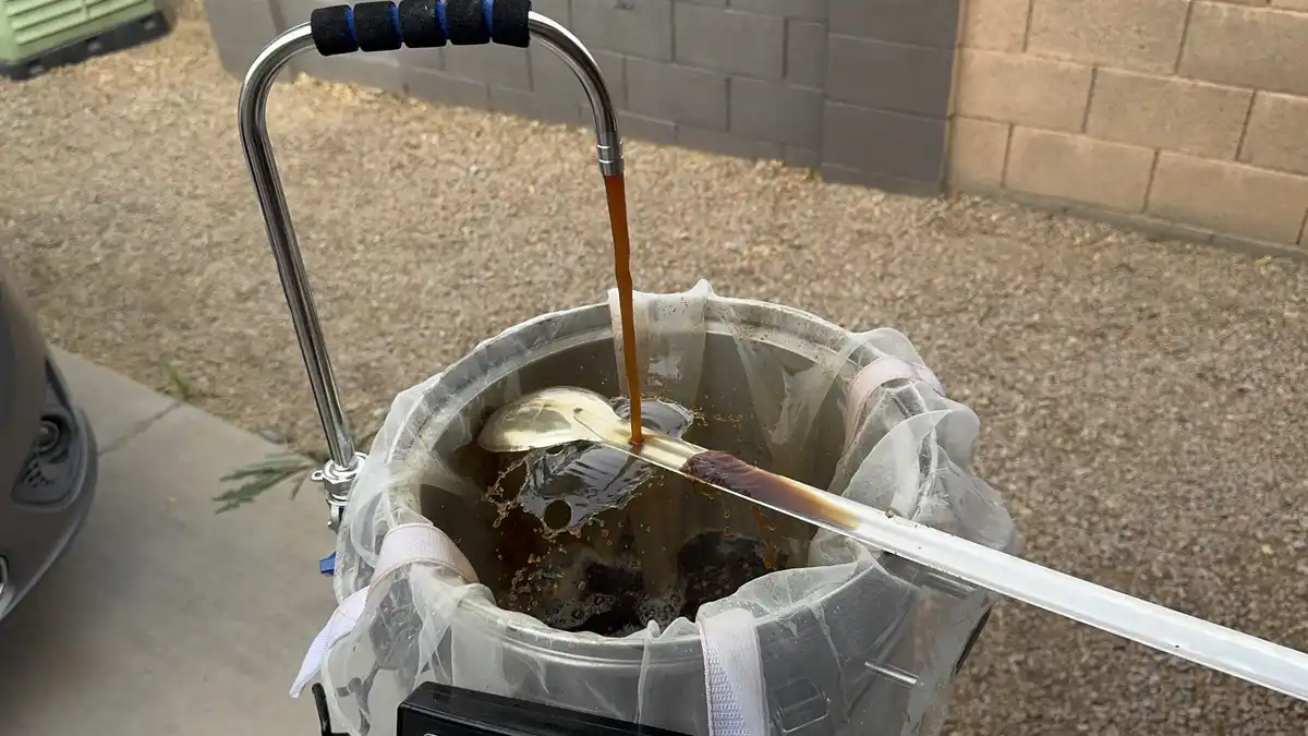A close-up shot shows dark coffee being poured from a metal spigot into a large metal bucket lined with a white mesh filter…