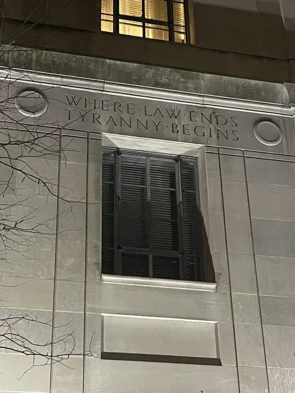 A nighttime photo of a stone building facade with engraved text reading 'WHERE LAW ENDS TYRANNY BEGINS' above a window with…