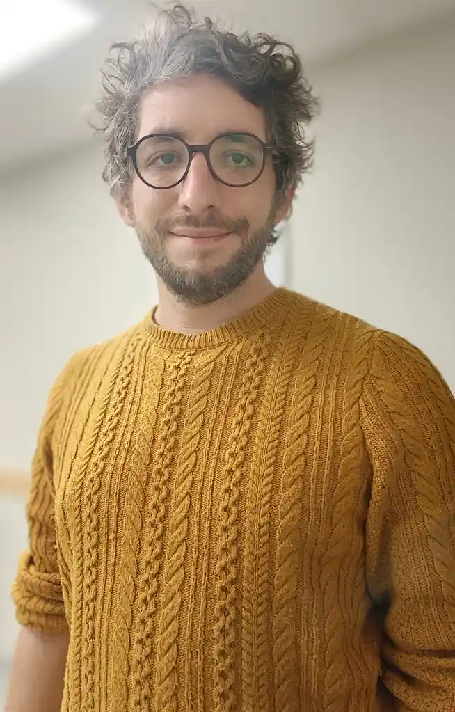 A man with curly grayish-brown hair, glasses, and a beard wears a mustard-yellow cable-knit sweater. He has a slight smile…