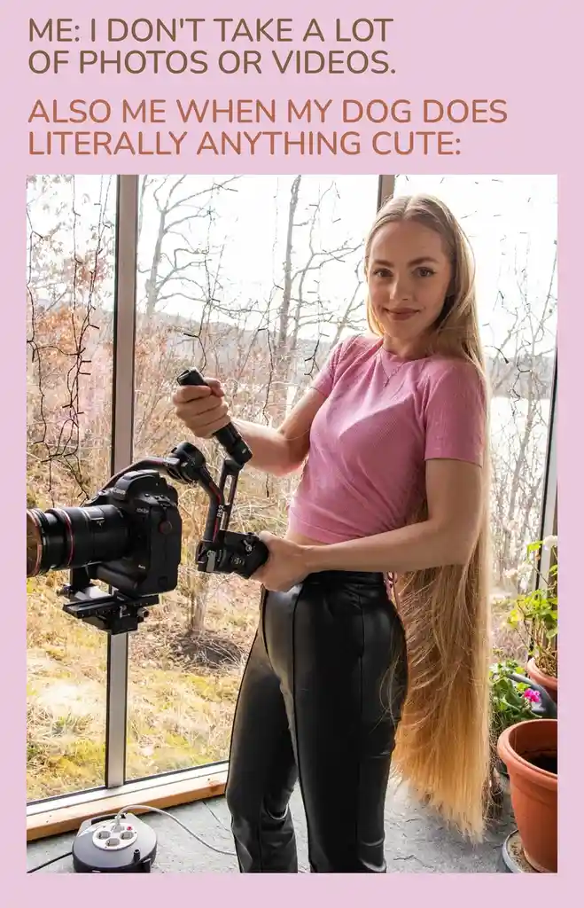 A woman with long blonde hair, wearing a pink shirt and black pants, stands indoors holding a professional camera mounted on…
