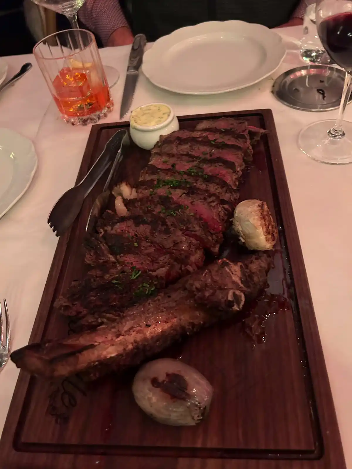 A large, sliced, medium-rare steak with a bone rests on a wooden serving board, garnished with herbs and accompanied by…