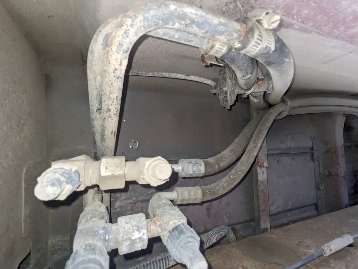A close-up photo of grimy, rusted, and dusty plumbing pipes and fittings inside a vehicle or machinery, with visible bolts…