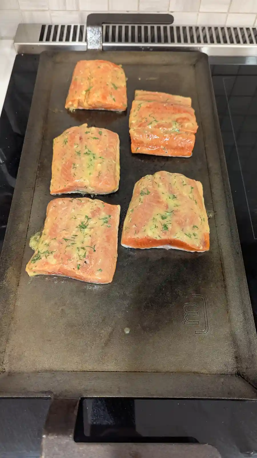 Five salmon fillets, seasoned with herbs and butter, are sizzling on a dark, well-used electric griddle. The fish is…