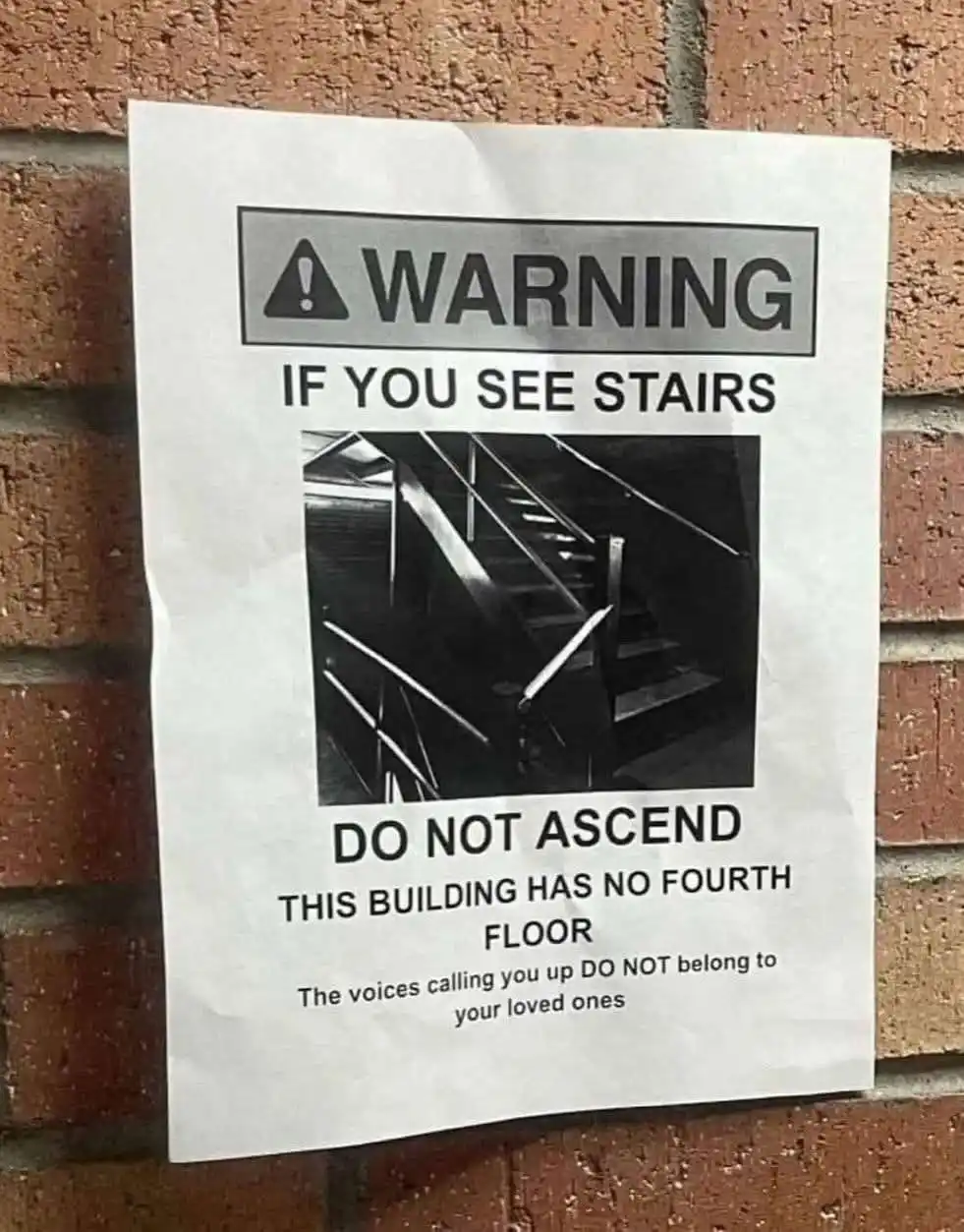 A crumpled warning sign taped to a brick wall features a black-and-white photo of a staircase. The text warns viewers not to…