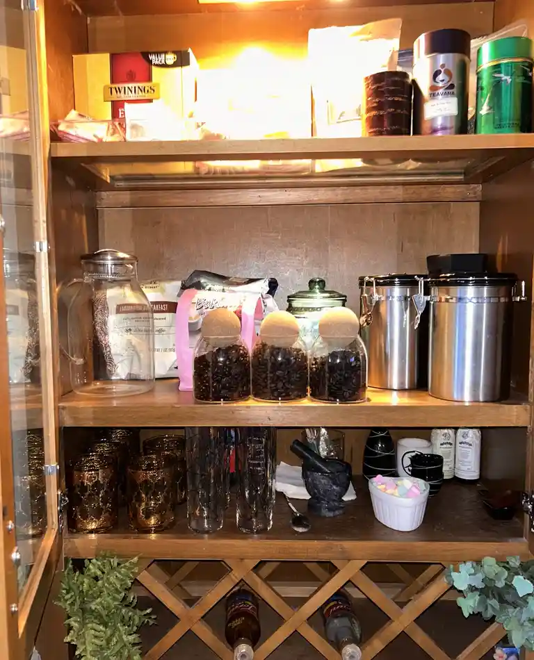 A well-lit wooden cabinet with glass doors displays three shelves filled with tea and coffee supplies. The top shelf holds…