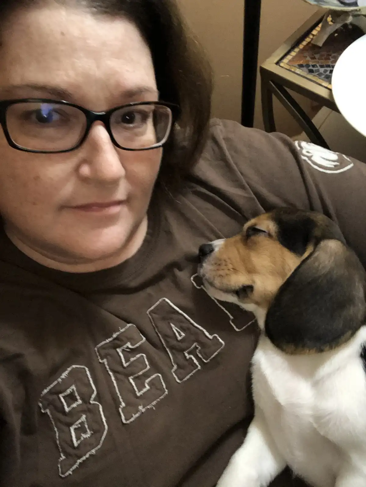 A woman wearing glasses and a brown shirt with the word 'BEA' visible holds a sleeping beagle puppy on her shoulder. The…
