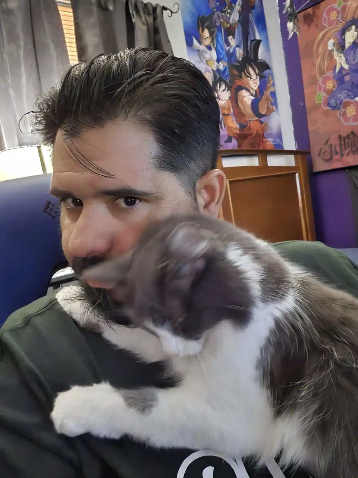 A man with dark hair and a mustache looks directly at the camera with a stern expression while holding a fluffy gray and…