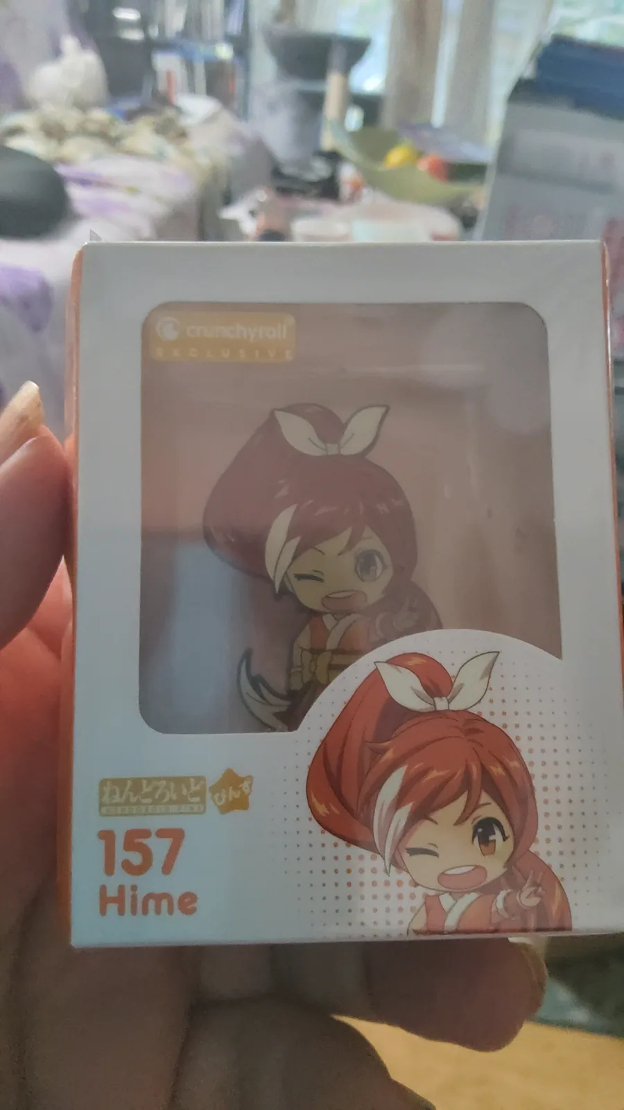 A hand holds a white box containing a chibi-style anime figurine of a character with red hair tied in a high ponytail with a…