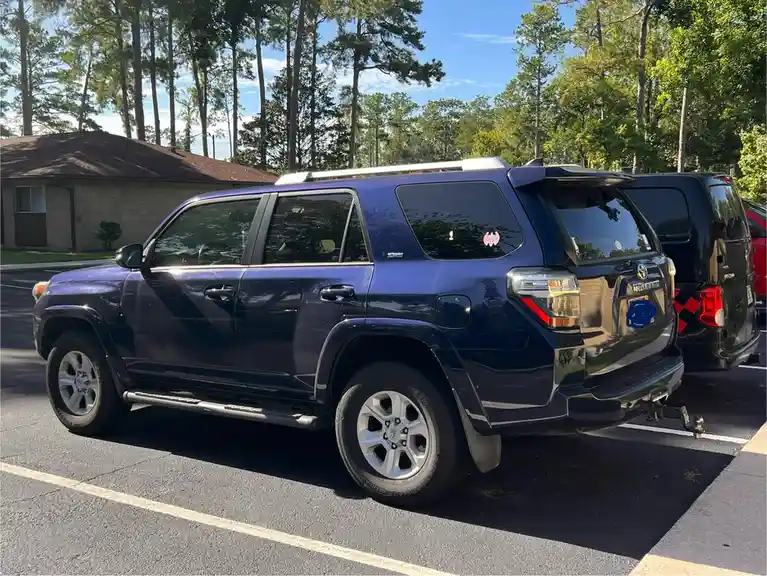 A dark blue Toyota 4Runner SUV is parked in a lot with trees and a house in the background. The rear license plate area is…