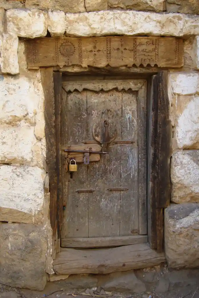 A weathered wooden door set in a stone wall is secured with a rusty metal bar and a small brass padlock, with faded graffiti…
