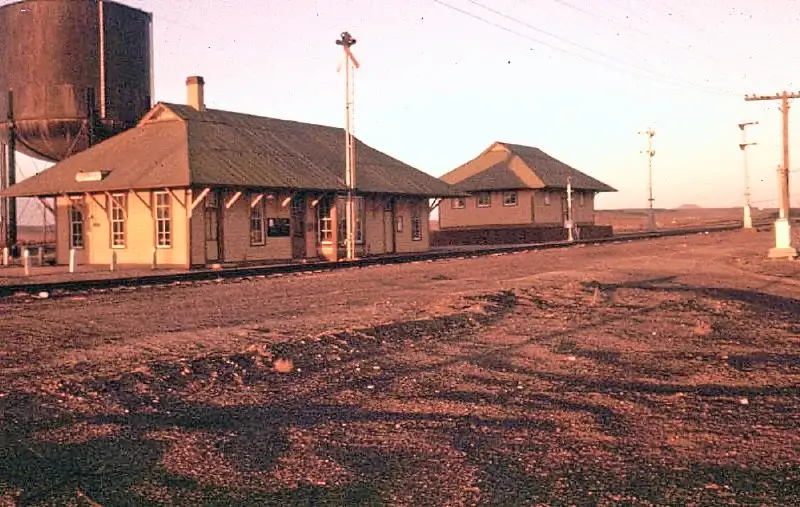 a sepia-toned photograph of a small, abandoned-looking train station with a single-story building, a water tower, and…