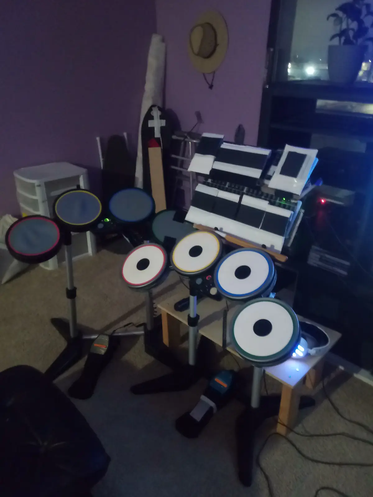A room with a purple wall and carpeted floor contains a large, complex electronic drum set with multiple colored pads…