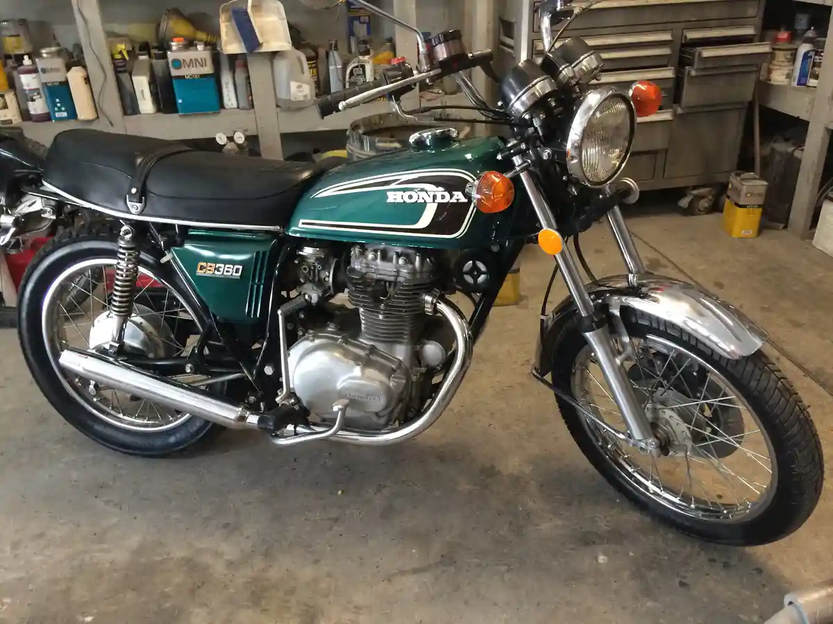 A green Honda CB360 motorcycle is parked in a cluttered garage with tools and supplies on shelves and a metal workbench in…