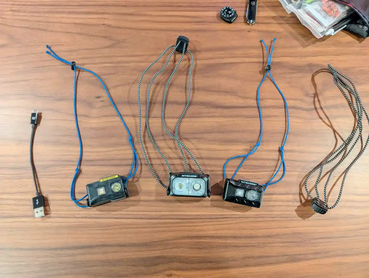 Three Nitecore brand headlamps with lanyards are laid out on a wooden surface, alongside a USB cable and other small gear…
