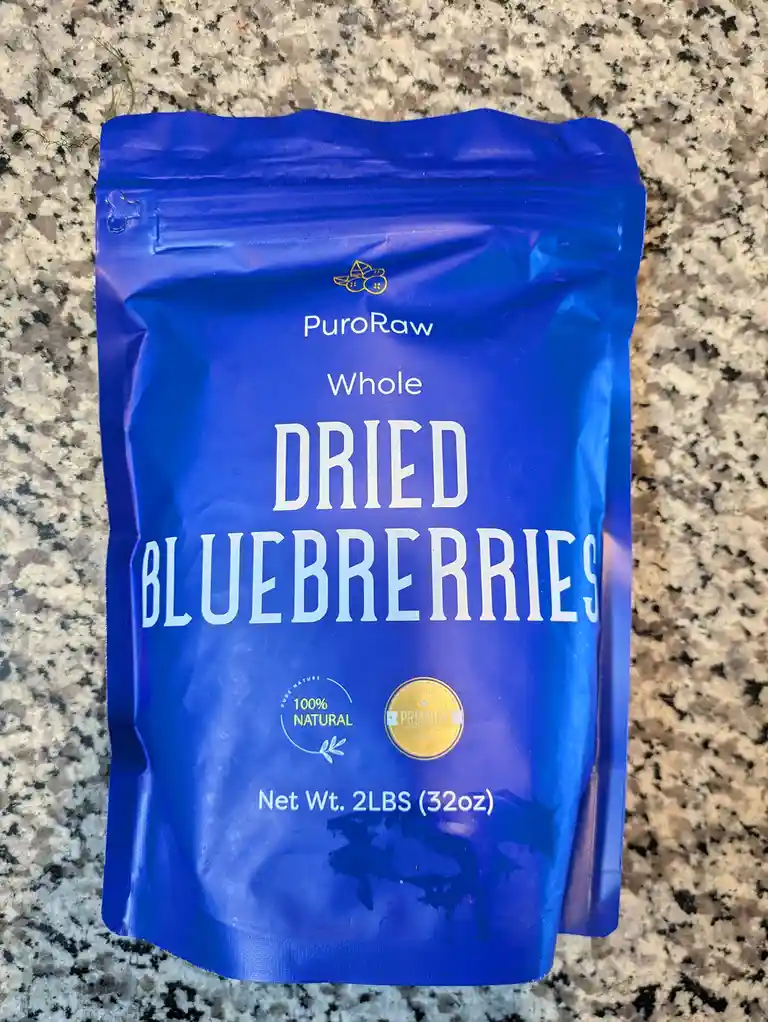 A blue resealable bag of PuroRaw Whole Dried Blueberries sits on a speckled granite countertop. The packaging prominently…
