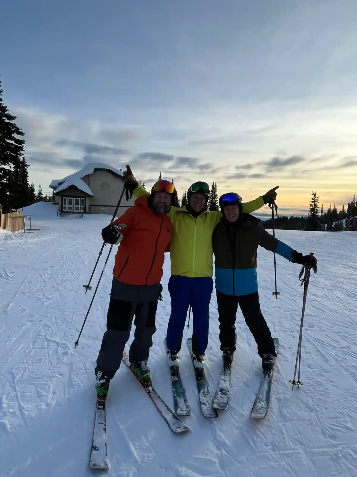 Three smiling skiers in full gear pose together on a snowy slope at sunset, arms around each other, with a ski lodge visible…