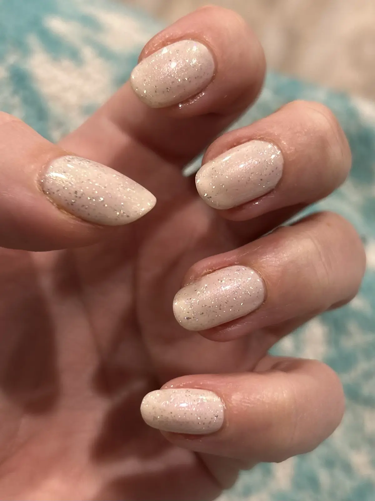 A close-up photo of a hand with five fingernails painted in a light pink, glittery polish. The nails are neatly manicured…