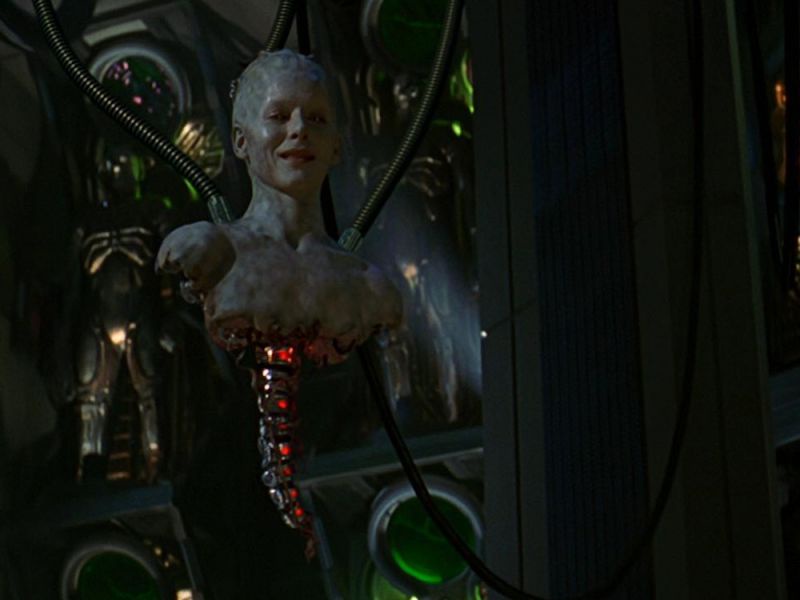 A pale, smiling humanoid torso with visible internal red glowing circuitry and tubes attached, suspended in a dark, sci-fi…