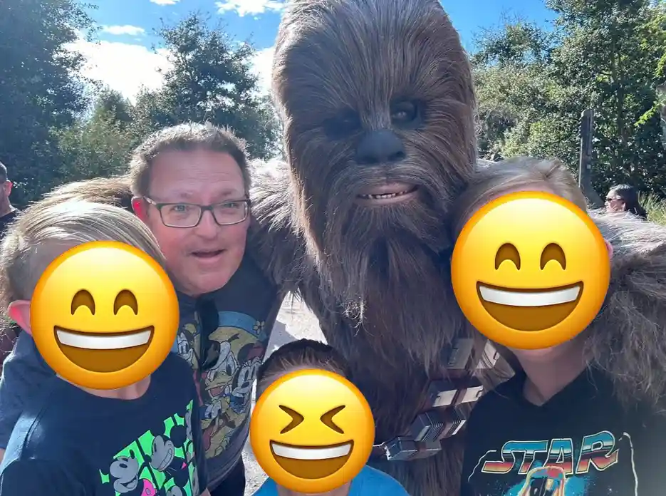 A family photo at a theme park features a person in a full Chewbacca costume standing with three children and an adult. The…