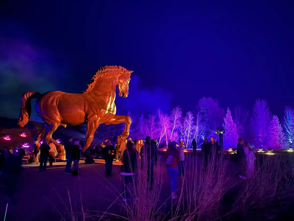 A large, golden horse statue stands illuminated at night, surrounded by people in a park with purple-lit trees. The overlaid…