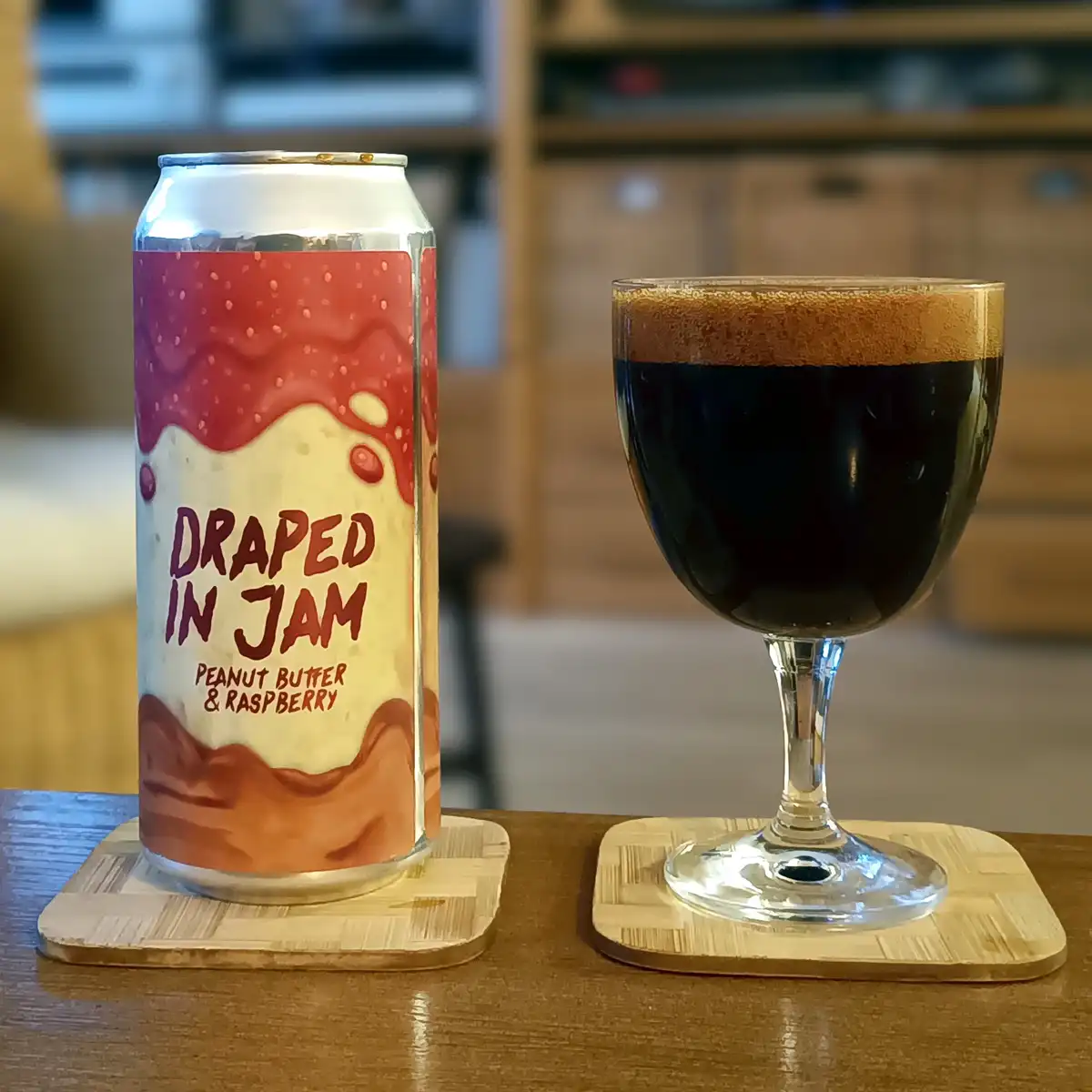 A beer can labeled 'Draped in Jam Peanut Butter & Raspberry' sits next to a glass of dark beer with a foamy head, both…