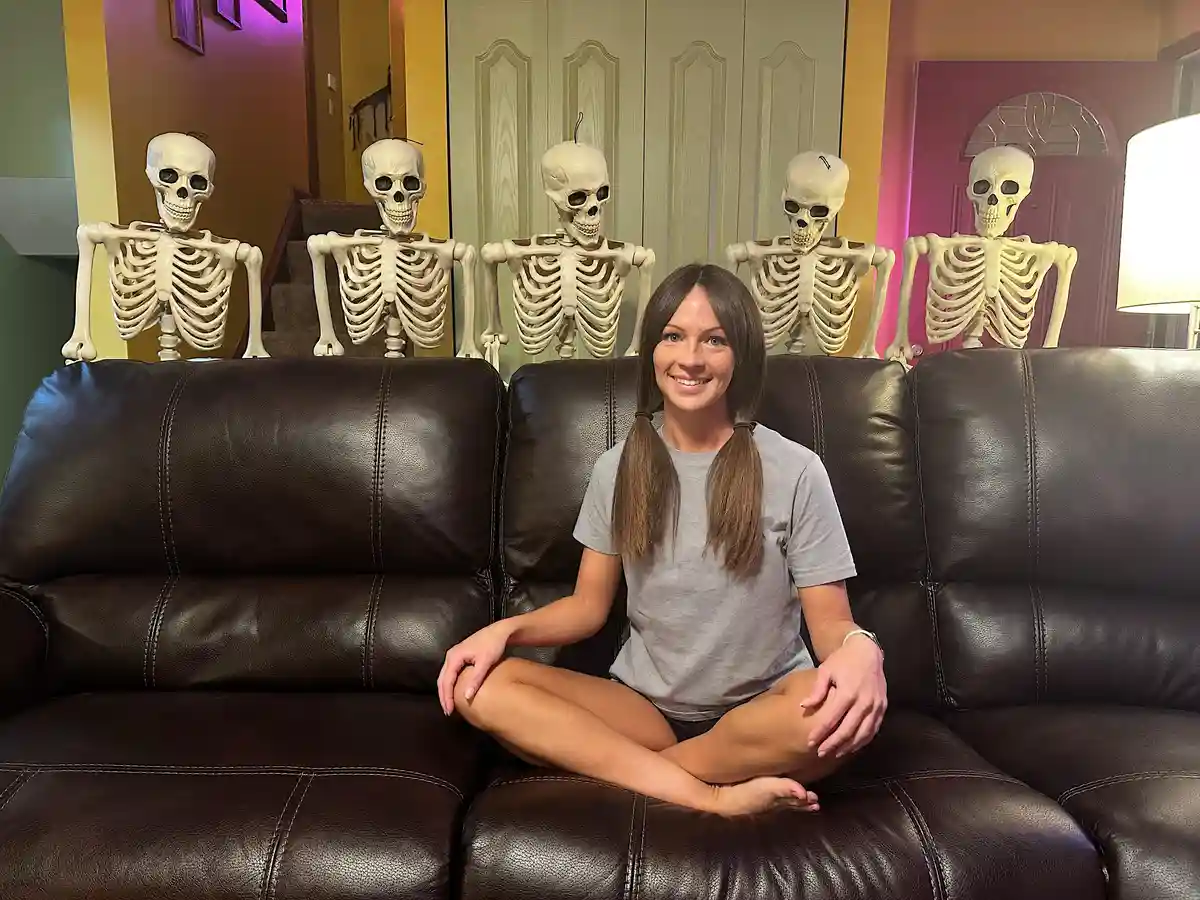 A woman sits cross-legged on a dark brown leather couch, smiling at the camera. Behind her, five full-sized plastic…