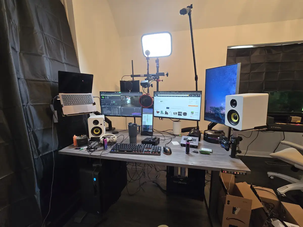 A cluttered home streaming or content creation desk features multiple monitors, a laptop, white speakers, a microphone, and…