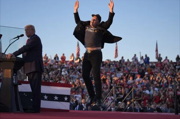 A man in a black cap and jacket, arms raised in celebration, stands on a stage next to Donald Trump at a rally. Trump stands…