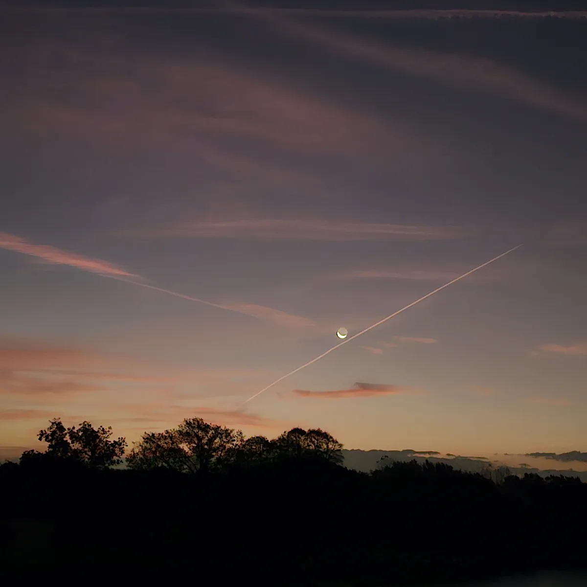 A crescent moon is visible in a twilight sky with pink and purple clouds, intersected by a long airplane contrail…