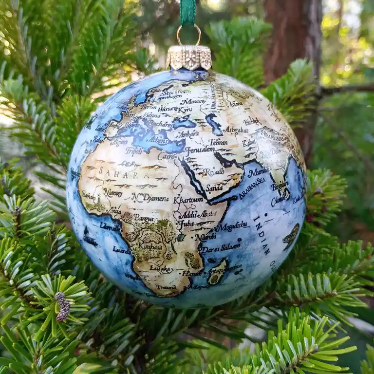A globe-shaped Christmas ornament with a vintage map of Africa, Europe, and Asia hangs from a green ribbon on a pine tree…