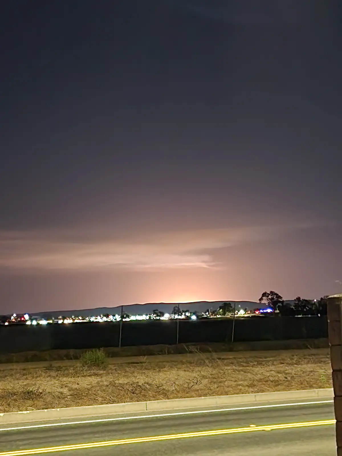 A nighttime photo shows a road with yellow lines, a fence, and distant city lights under a dark sky with a faint orange glow…