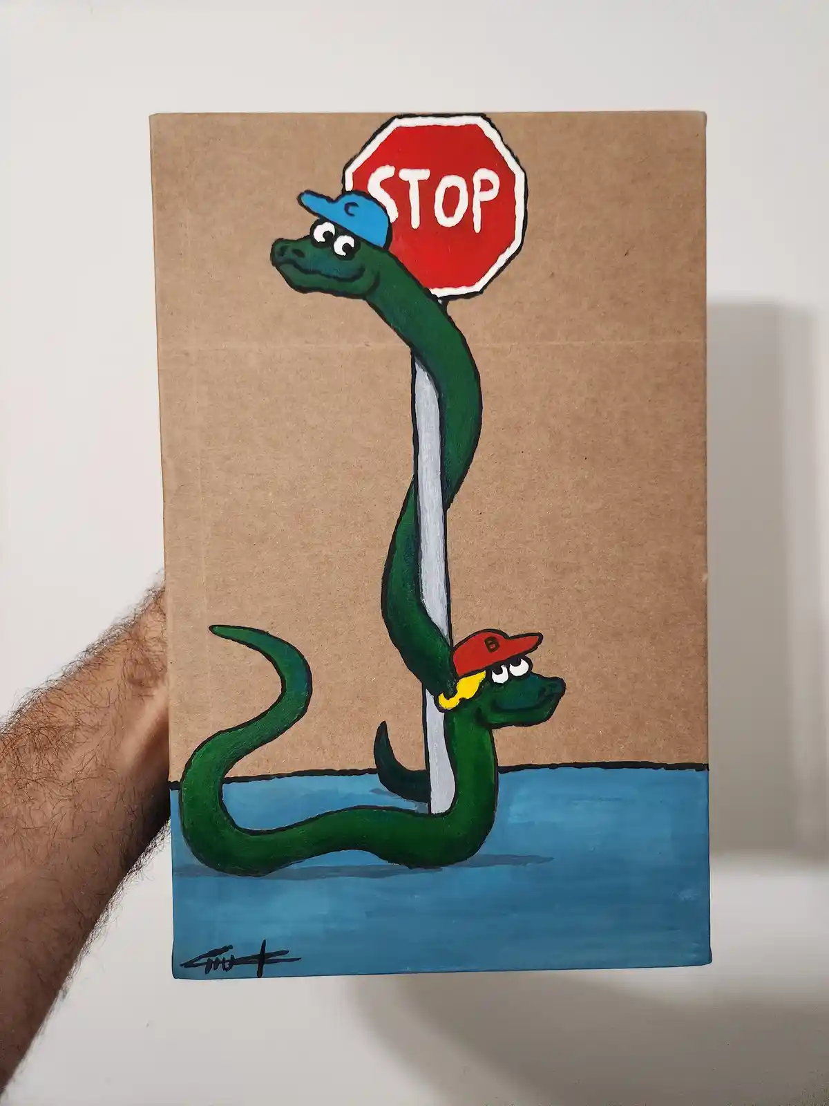 A hand holds a cardboard drawing showing two cartoon green snakes, one wearing a blue cap and holding a red stop sign, the…