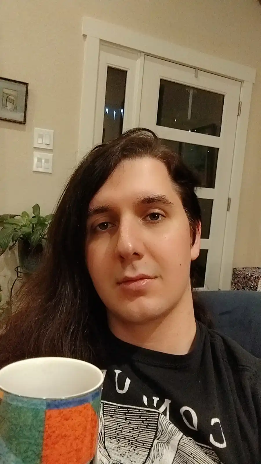 A person with long dark hair sits indoors holding a colorful mug, looking directly at the camera with a neutral expression…