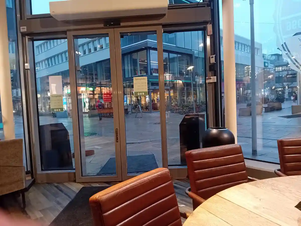 A photo taken from inside a cafe or shop looking out through automatic glass doors onto a modern urban plaza. The interior…