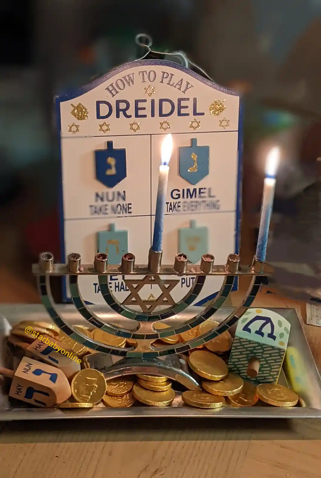 A menorah with two lit blue candles is placed in front of a 'How to Play Dreidel' guide, surrounded by gold coins and…