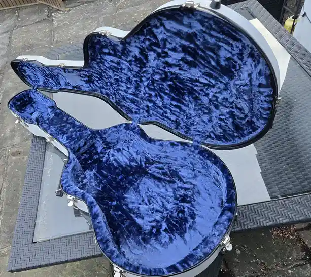 An open, silver-colored guitar case with plush blue velvet lining sits empty on a glass-topped outdoor table. The case is…