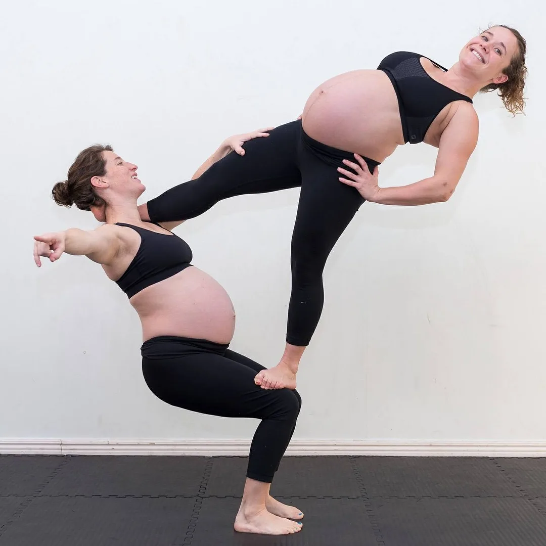 Two pregnant women in black workout attire perform a balancing yoga pose against a plain white wall; one woman is squatting…